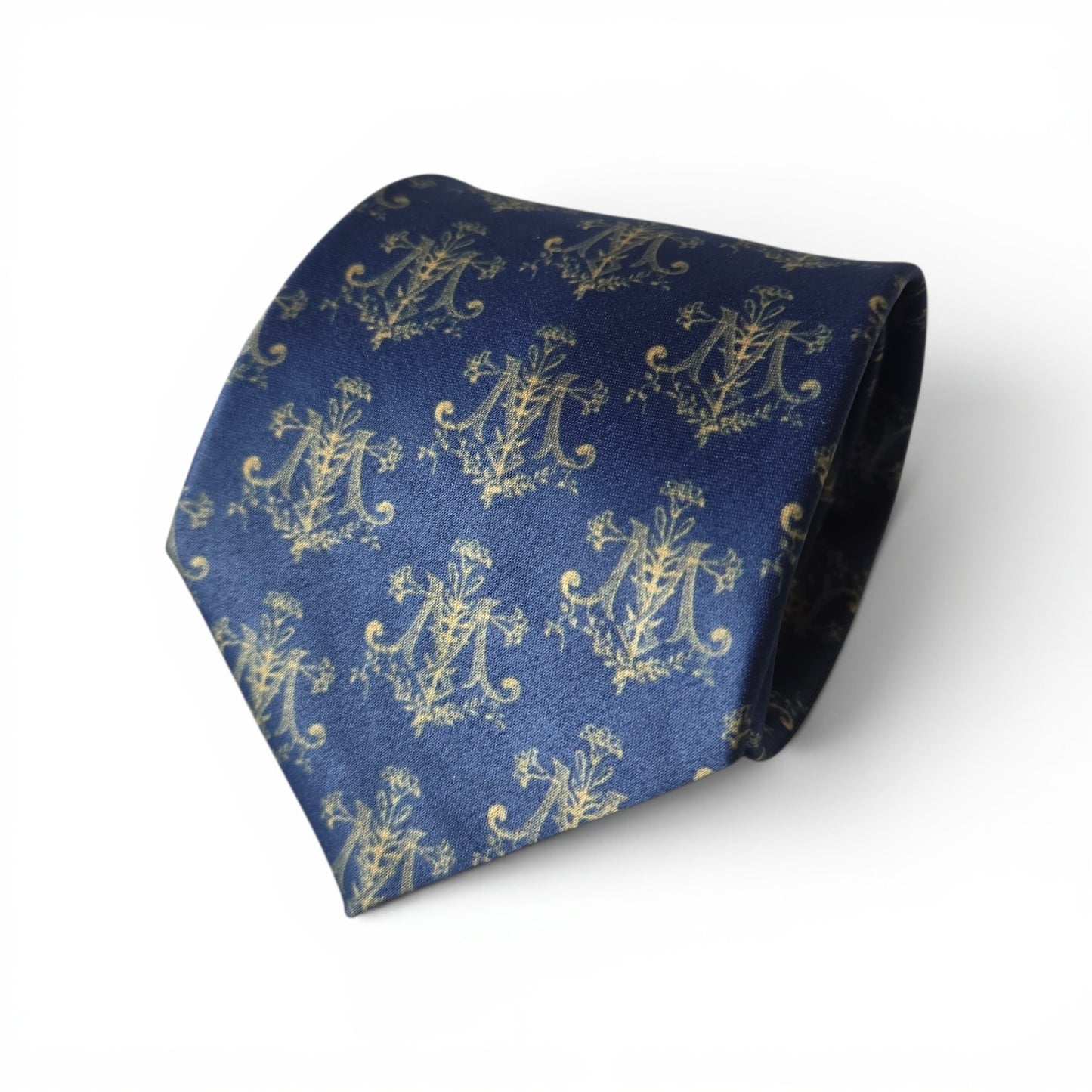 Auspice Maria necktie in navy blue with repeating gold ornate Marian monogram and floral pattern, rolled product shot on white background